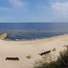 Plaża dla psów z łagodnym zejściem do morza. Na piasku ławki i kosze. W oddali widać falochron i statki.