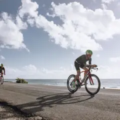 Dwóch kolarzy ściga się na trasie rowerowej wzdłuż Bałtyku. Słońce odbija się od wody, a chmury tworzą malowniczy krajobraz.