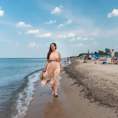 Kobieta w kwiecistej sukience spaceruje brzegiem morza. W tle plaża pełna ludzi, parawanów i namiotów – typowe sarbinowo atrakcje.