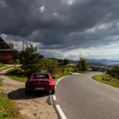 Czerwony kabriolet zaparkowany przy drewnianym domu. Droga prowadzi przez malownicze wzgórza, idealne miejsca, co zwiedzić po drodze do Zakopanego.