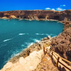 Fuerteventura na co uważać? Klify i turkusowe morze, ścieżka z drewnianą balustradą prowadzi w dół. Uważaj na nierówny teren.
