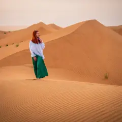 Kobieta w białej bluzce i zielonej spódnicy podziwia bezkresne wydmy. To jej przygoda, **Oman na własną rękę**.