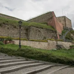 Kamienne schody prowadzą do potężnej twierdzy, jednej z głównych atrakcji Kłodzka. Widać metalowe schody i zielone zbocza.