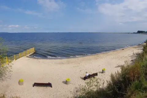 Plaża dla psów z łagodnym zejściem do morza. Na piasku ławki i kosze. W oddali widać falochron i statki.