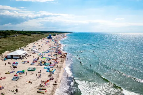 Słoneczna plaża w Darłowie tętni życiem. Tłumy plażowiczów, kolorowe parawany i namioty, a w wodzie kąpielowicze i surferzy. To idealne miejsce na wakacyjne atrakcje.