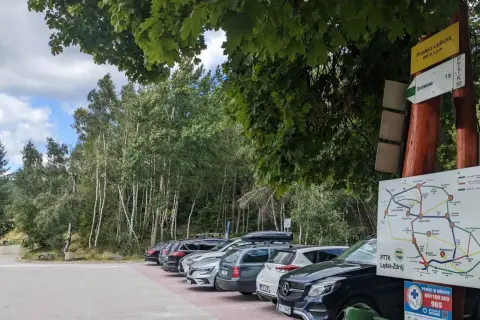Samochody zaparkowane przy szlaku turystycznym na przełęczy kłodzkiej. W tle las i tablice informacyjne PTTK.