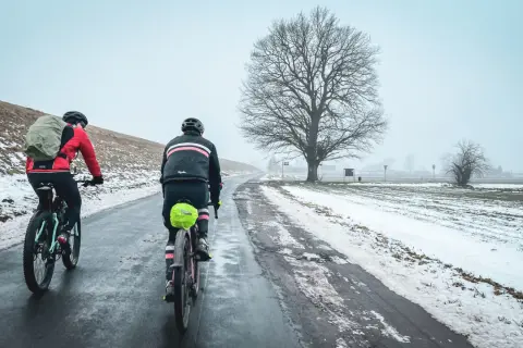 Dwaj rowerzyści na trasie Wisła 1200, w zimowej scenerii, pokonują oblodzoną drogę pod rozłożystym drzewem.