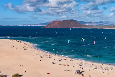 Fuerteventura, gdzie najlepiej? Plaża z kitesurferami i turystami, w tle wulkan i góry. Idealne miejsce na wakacje.