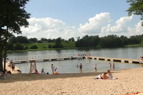 Letnie popołudnie na piaszczystej plaży nad jeziorem. Ludzie odpoczywają, kąpią się, a dzieci bawią się w wodzie. To typowy widok dla mazurskich miejscowości.