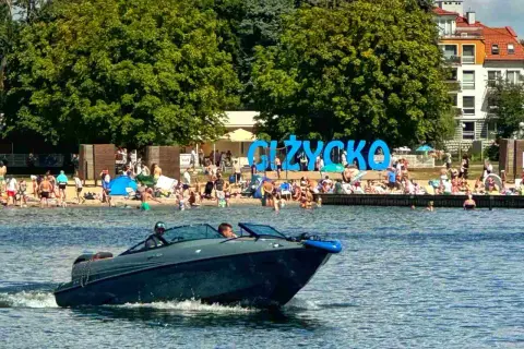 Łódź motorowa na jeziorze, plaża pełna ludzi i wielki napis GIŻYCKO. Idealne miejsce, gdzie na Mazury!