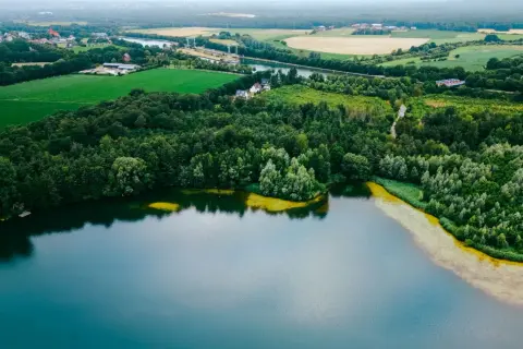 Malowniczy krajobraz z lasem i polami otaczającymi najczystsze jezioro w Polsce. W oddali widać zabudowania i kanał.