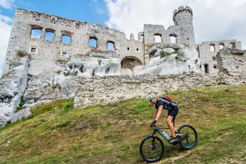 Rowerzysta pokonuje wzgórze na Szlaku Orlich Gniazd, w tle ruiny zamku.
