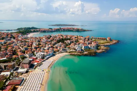 Malownicze wybrzeże Bułgarii nad turkusowym morzem. Miasto z czerwonymi dachami rozciąga się wzdłuż piaszczystej plaży z białymi parasolami.