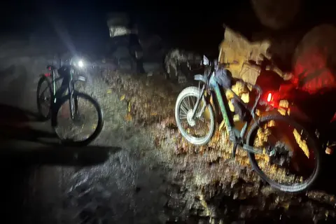 Dwa rowery z włączonymi światłami stoją na wilgotnej ścieżce. Nocna wyprawa po trasach rowerowych Elbląg.
