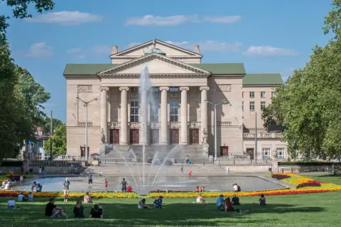 Poznań atrakcje dla dorosłych: Teatr Wielki i fontanny w słoneczny dzień. Ludzie odpoczywają na trawie, ciesząc się miejskim krajobrazem.