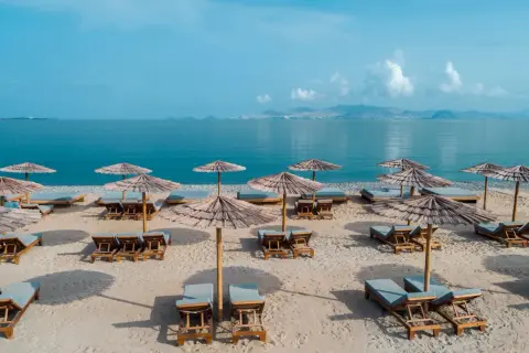 Puste leżaki i parasole na piaszczystej plaży, gotowe na gości. W tle spokojne, błękitne morze i górzyste wybrzeże. Idealne miejsce na relaks.