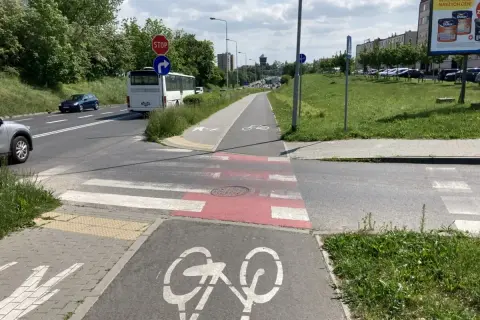 Pieszy na ścieżce rowerowej czeka na zielone światło, by przejść przez pasy. Obok droga z autobusem i samochodami.