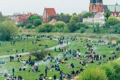 Ludzie odpoczywają na trawie w nadwarciańskiej części Poznania. W tle widać zabytkowe budynki i rzekę.