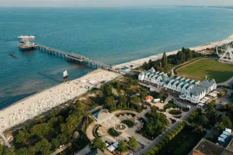 Widok na plażę, molo i białe budynki z dachami w szpic na wyspie Uznam. W oddali widać diabelski młyn.