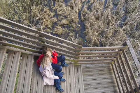 Dwoje dzieci siedzi na drewnianych schodach przy bagnistym terenie. Starsza dziewczynka przytula młodszą, jakby to było ich pierwsze bagno całowanie.