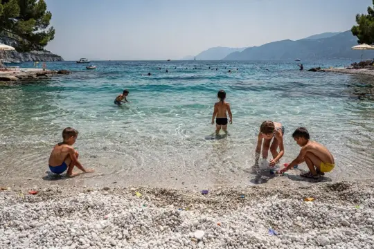 Dzieci bawią się w płytkiej, krystalicznie czystej wodzie na kamienistej plaży. W tle widać góry i łodzie. To idealne plaże w Chorwacji na rodzinne wakacje.