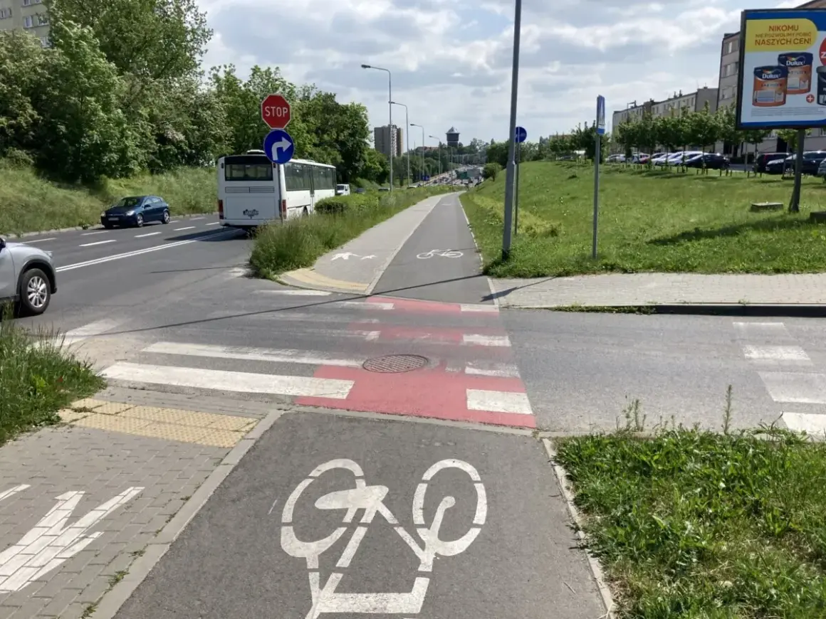 Pieszy na ścieżce rowerowej czeka na zielone światło, by przejść przez pasy. Obok droga z autobusem i samochodami.