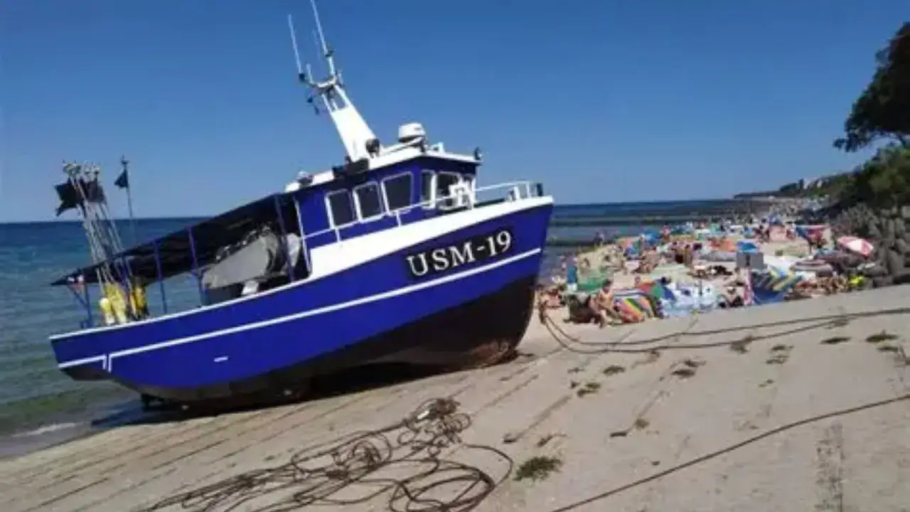 Niebiesko-biała łódź rybacka USM-19 na plaży, w tle tłum plażowiczów. Ustronie Morskie atrakcje w pełnym słońcu.