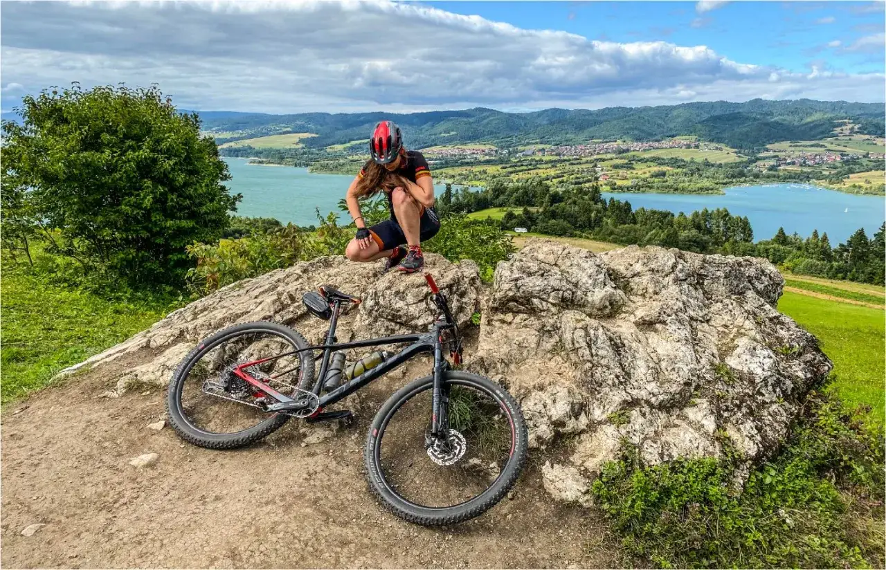 Kobieta w kasku odpoczywa na skale obok roweru, podziwiając malowniczy widok na jezioro i góry. Idealne miejsce na odkrywanie velo tras rowerowych.
