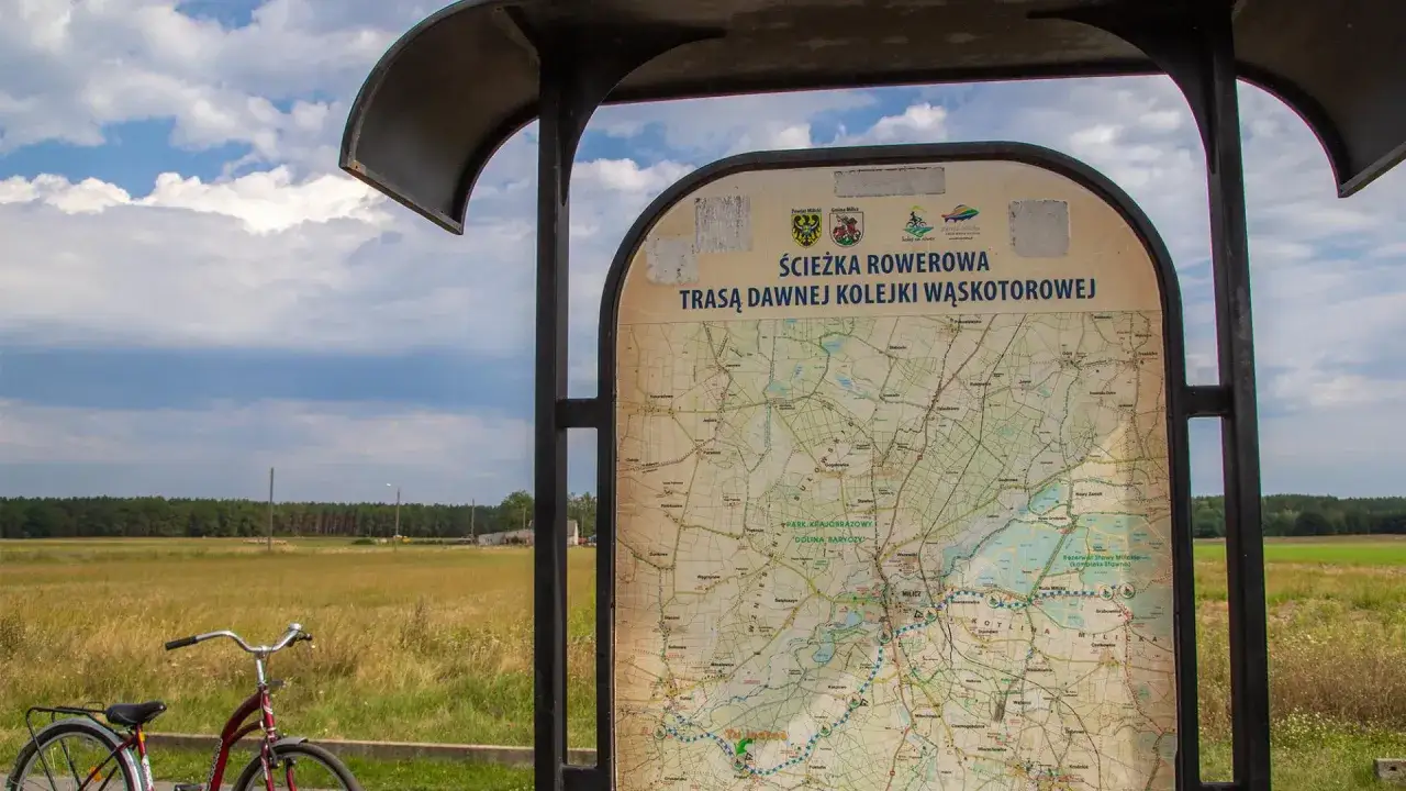 Mapa turystyczna informuje o ścieżce rowerowej trasą dawnej kolejki wąskotorowej. Obok stoi rower.