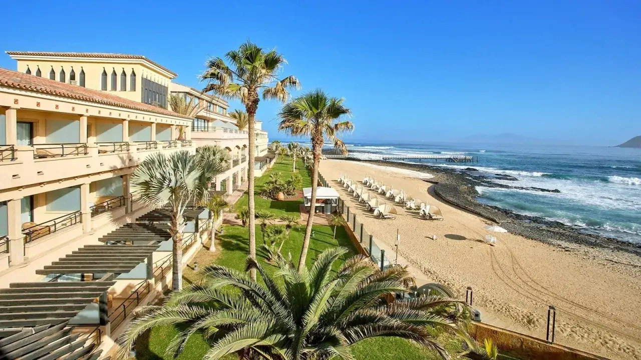 Hotel z palmami przy plaży, idealne miejsce na Fuerteventurze, gdzie najmniej wieje. Spokojne fale oceanu i złoty piasek.