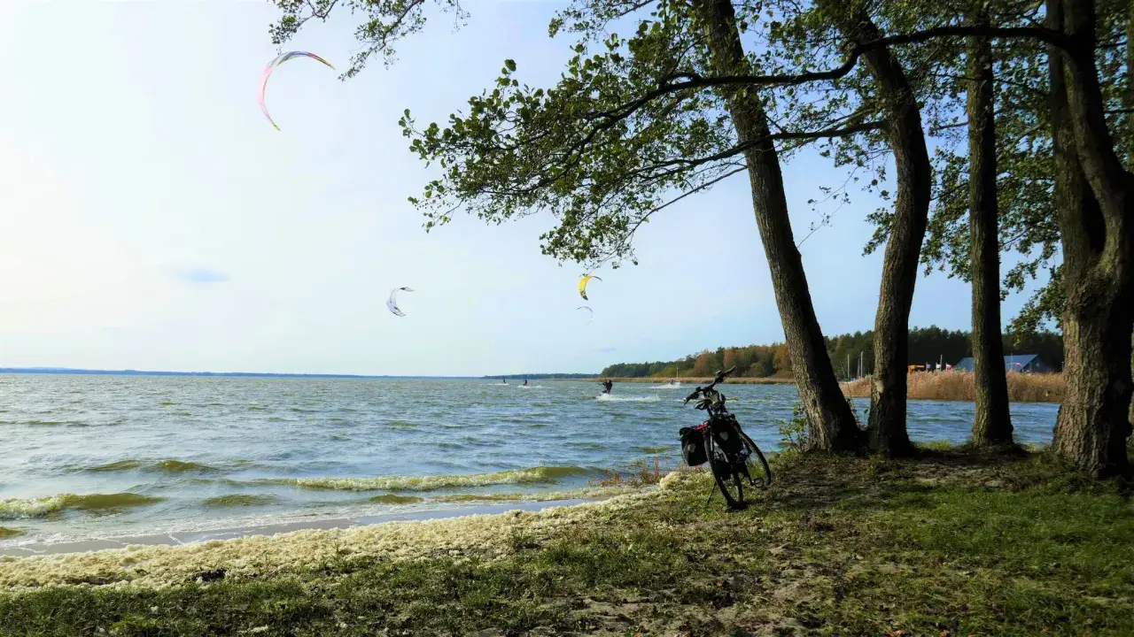 Dąbki atrakcje: kitesurferzy na jeziorze, rower przy drzewach, piana na brzegu.