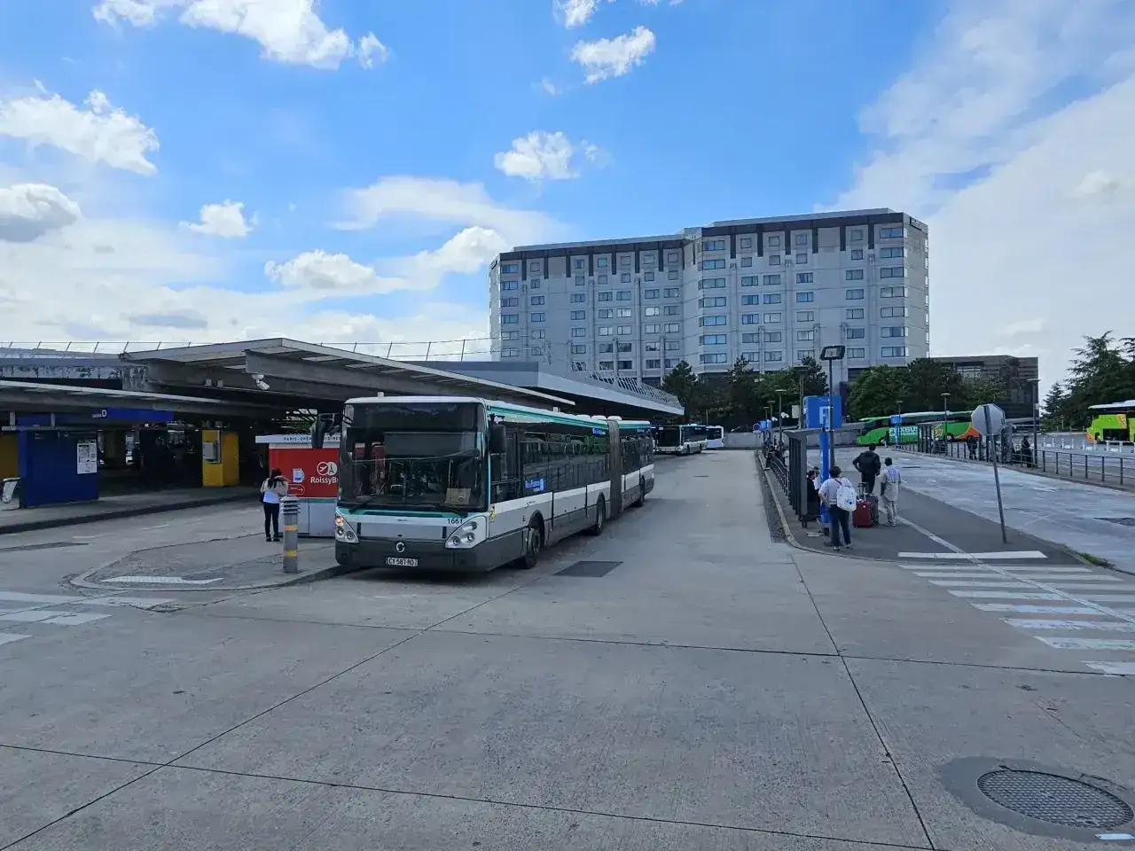 Autobusy czekają na pasażerów na przystanku, który jest częścią infrastruktury lotniska w Paryżu. W tle widać hotel i inne budynki.
