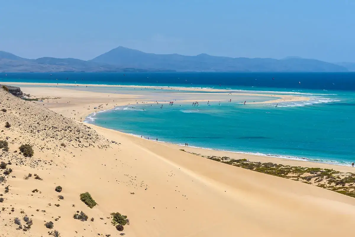 Plaża na wyspie Fuerteventura z turkusową wodą, piaskami i licznymi surferami.