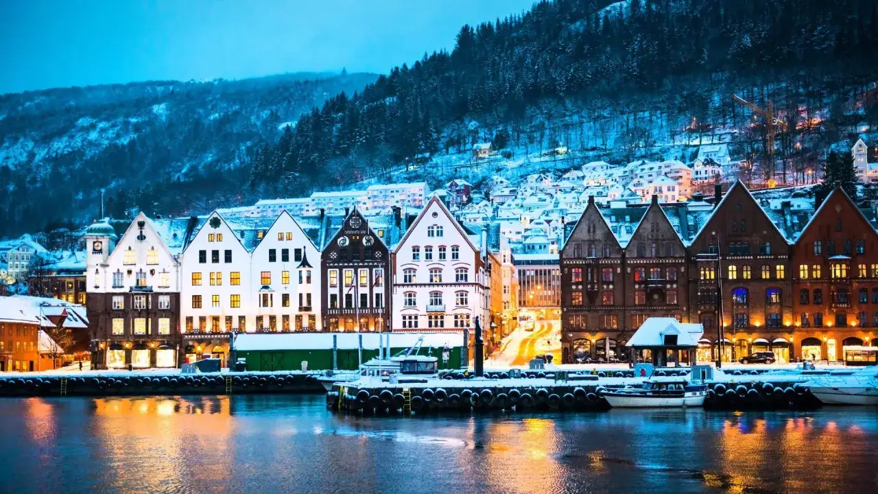 Zimowy wieczór w Bergen, kiedy najlepiej jechać do Norwegii? Kolorowe kamienice nad wodą, ośnieżone wzgórza w tle.